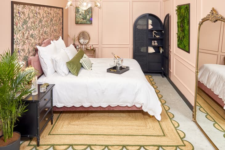 Side view of a pink velvet bed frame with white scalloped duvet. The room has pink walls, a black armoire and side table, and a gold framed standing mirror.