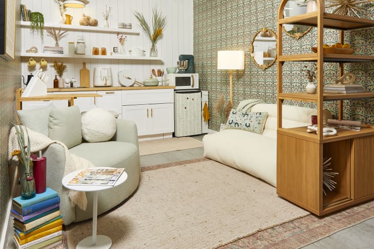 Head on view of a small living room with sage and light pink floral wallpaper on the right wall and white wood panels on the left. The kitchen has white cabinetry and there a large cream colored chair on the right and a light green swivel chair on the left.