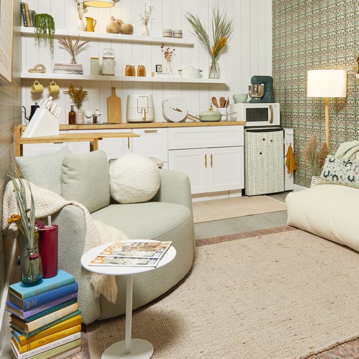 Head on view of a small living room with sage and light pink floral wallpaper on the right wall and white wood panels on the left. The kitchen has white cabinetry and there a large cream colored chair on the right and a light green swivel chair on the left.