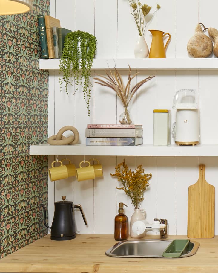 Close up view of white open shelving against a white wood paneled wall. The shelves have assorted books, plants and kitchen accessories.