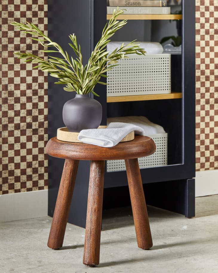 Angled view of a small wooden stool with a small beige tray, a white towel and a small black vase with olive branches in it.