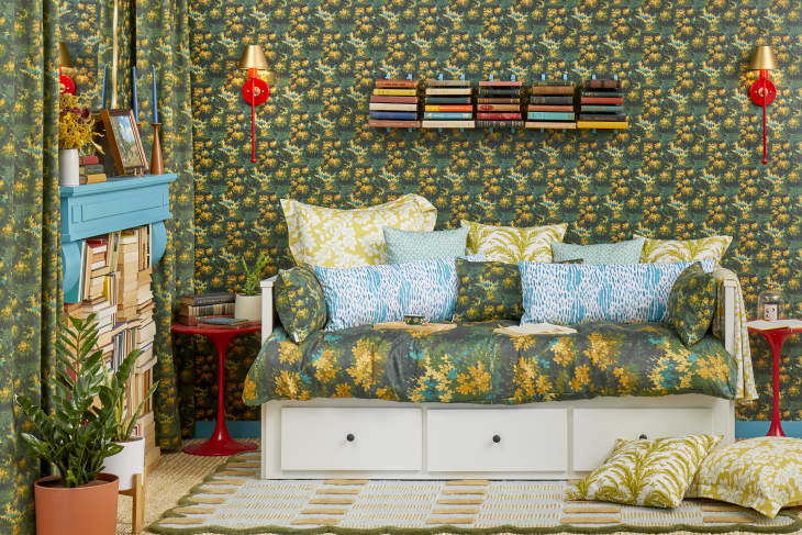 Head on view of a white day bed, in a room with green and yellow floral wallpaper.  The bed is made with a large assortment of pillows with different prints.