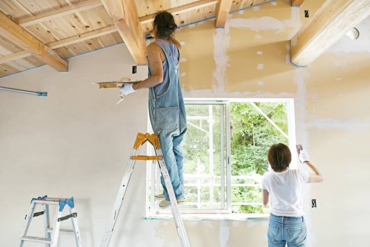 2 people, one on a ladder, painting the walls of a house white. The ceiling is exposed wood. There is a window letting in daylight and trees are outside