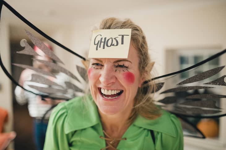 Woman dressed as a fairy laughing while wearing a sticky note on her head that says 'ghost' as she plays a game at a Halloween party.