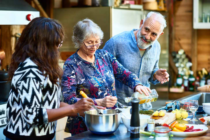 Woman wearing glasses pouring oil and making food, mature man with beard talking to female friend, working as a team, three people cooking at home.