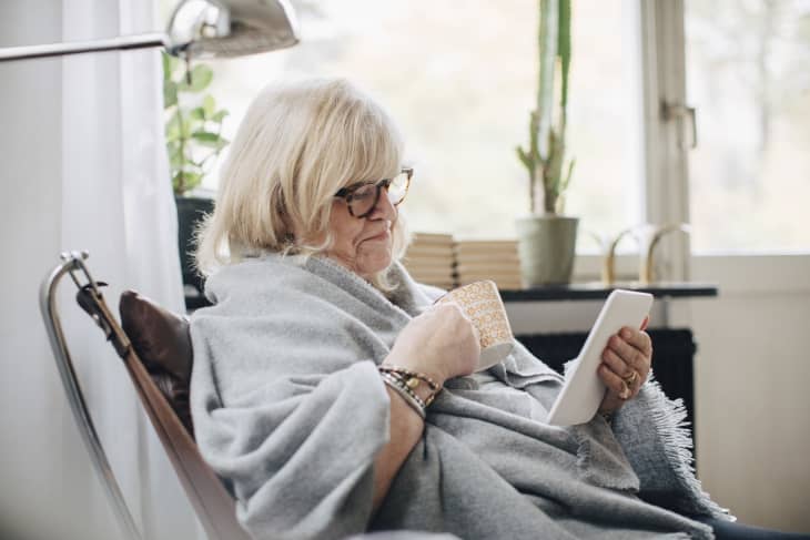 Retired senior woman wrapped in blanket holding coffee cup reading e-book in room at home