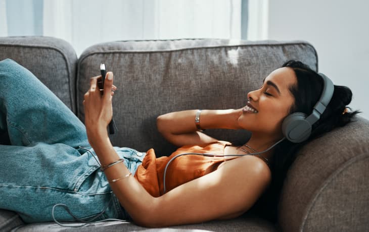 一个年轻女子坐在家里的沙发上使用智能手机和耳机