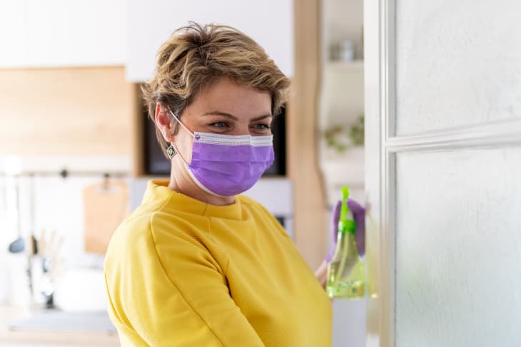 Woman with protective mask and gloves using disinfectant and rag to clean glass door