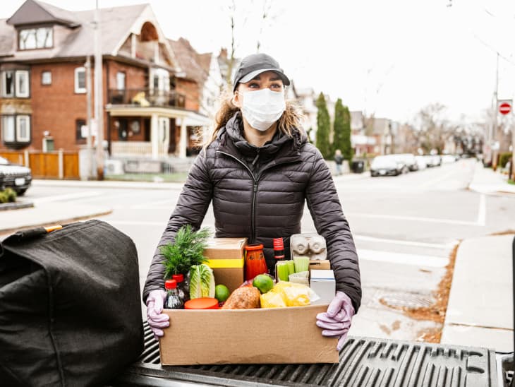 COVID-19, Young women delivers fresh food during pandemic lockdown. She is offloading box of fresh produce from the bed of the pick up truck. She is wearing face mask. Urban setting of a North American city.