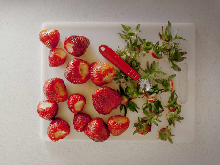 White cutting board with hulled strawberries, their tops, and the strawberry hulling tool
