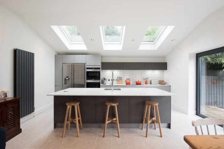 A general view of a modern fitted grey kitchen with large island, breakfast bar, white walls and sky light windows within a home