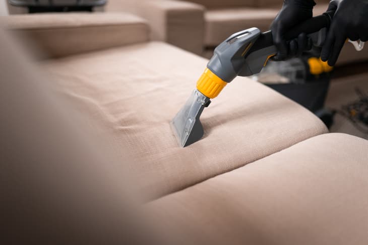 Sofa cleaning by a device. Close-up of housekeeper hand holding modern washing vacuum cleaner and cleaning dirty sofa with professionally detergent.