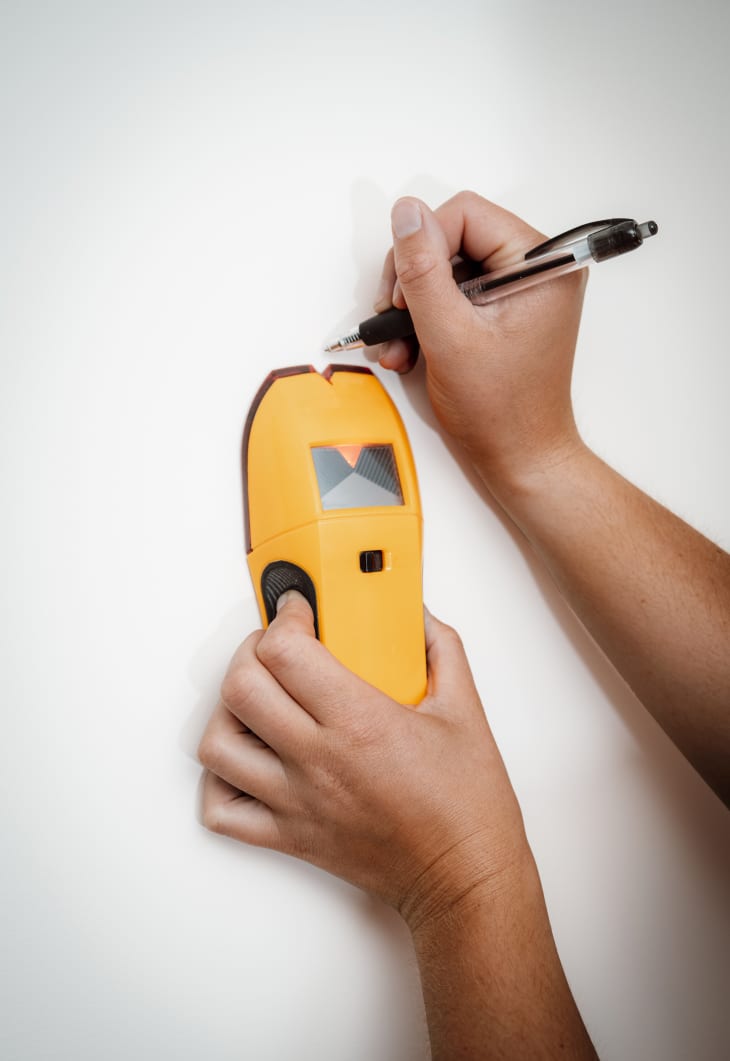 hands holding and using stud finder on a white wall, marking with a pen above