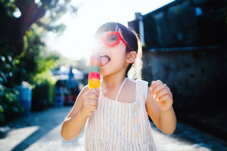 Happy little girl with red sunglasses eating colourful popsicle outdoors on a hot summer day, standing against nature scene with trees and sky