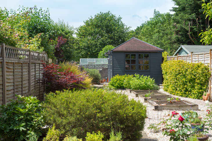 A view looking down a back garden of a home with gravel, pea shingle, wooden railway sleeper flower beds, vegetable patch, potted plants, timber fence and grey summerhouse, shed with tiled roof on a warm sunny day