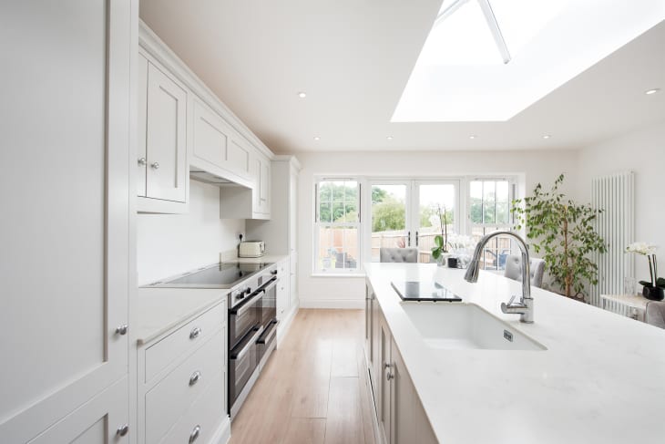 A general interior view of a domestic dove grey kitchen, with painted white walls, island with quartz worktop, chrome tap, grey crushed velvet bar stools, stainless steel range cooker oven, chrome tap, skylight, vertical radiator, potted plant and patio doors into the back garden within a home