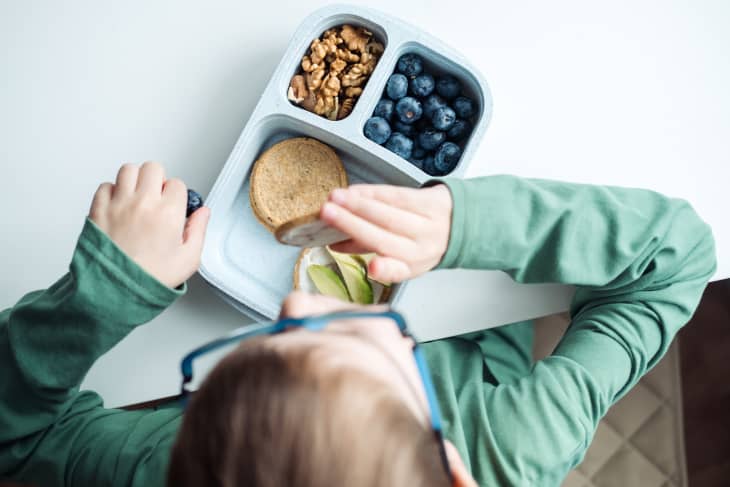 A schoolboy in glasses eats from a lunchbox, avocado, cracker, cheese, blueberries and walnuts while sitting at the table, top view