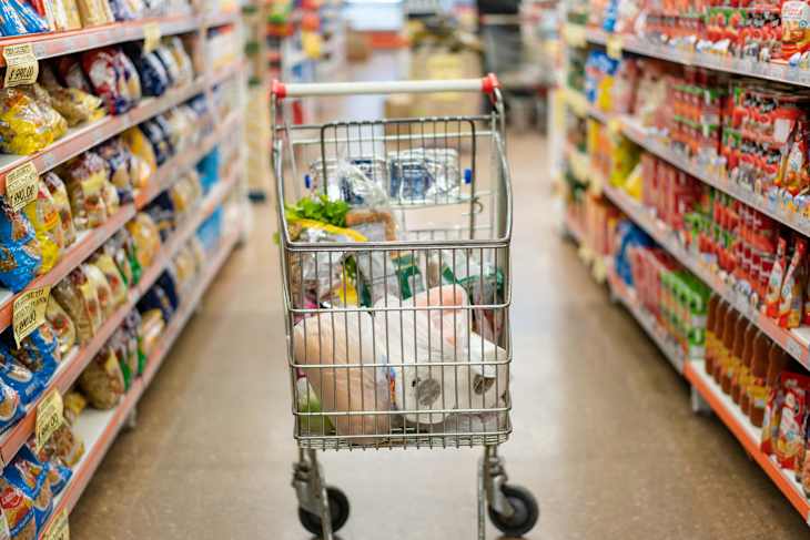 Shopping cart with products in the middle of supermarket aisle. Shopping cart with heap of various groceries along the shelves at modern grocery store.