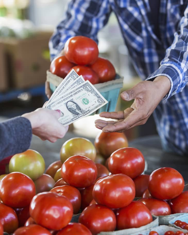 Cropped view of a worker at a produce stand taking cash from a customer. She is handing over paper currency. Fresh, ripe tomatoes are on the table between them.