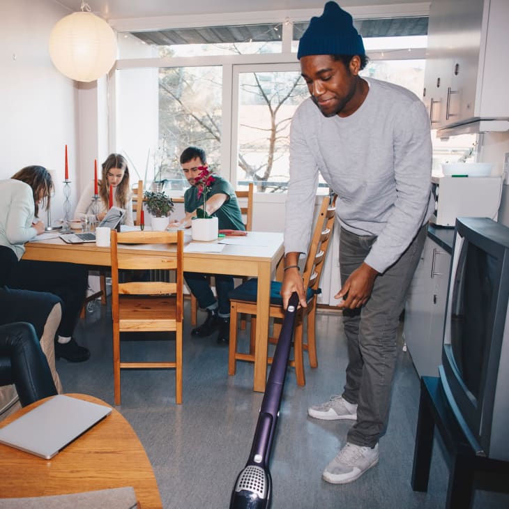 Man cleaning floor while friends sitting in college dorm room