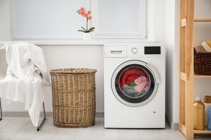 A photo of a washing machine with clothes inside, a circular laundry hamper, and a chair with clothes hang on it
