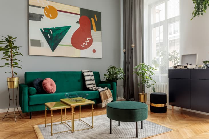 Luxury and modern home interior with design green sofa, navy commode, tables, pouf and accessroies. A lot of plants in the room. Abstract painting.  Stylish decor of living room with brown parquet.