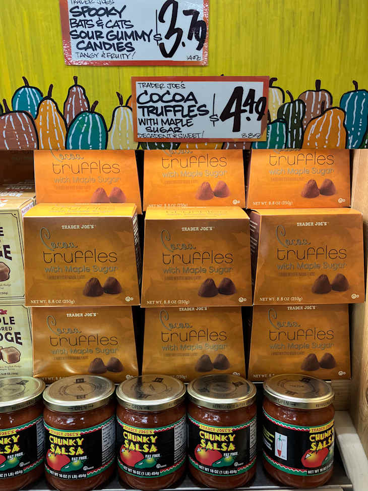 Trader Joe's Cocoa Truffles with Maple Sugar stacked on shelves, alongside jars of Chunky Salsa. Colorful pumpkin-themed sign above.