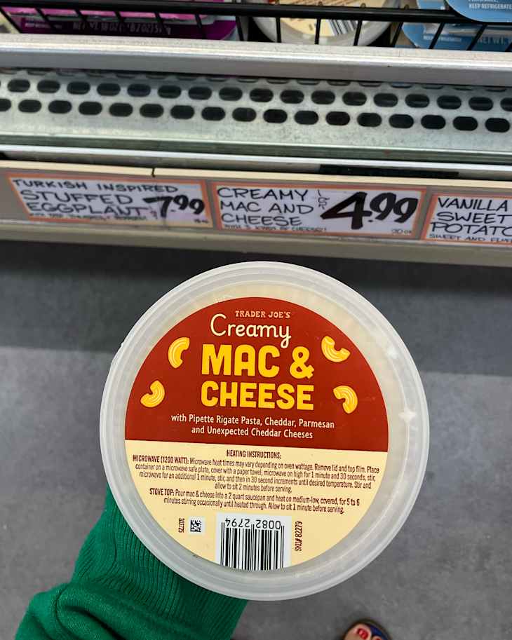 Creamy mac and cheese container from Trader Joe's held by a person wearing a green sweater, with price tags for other products in the background.