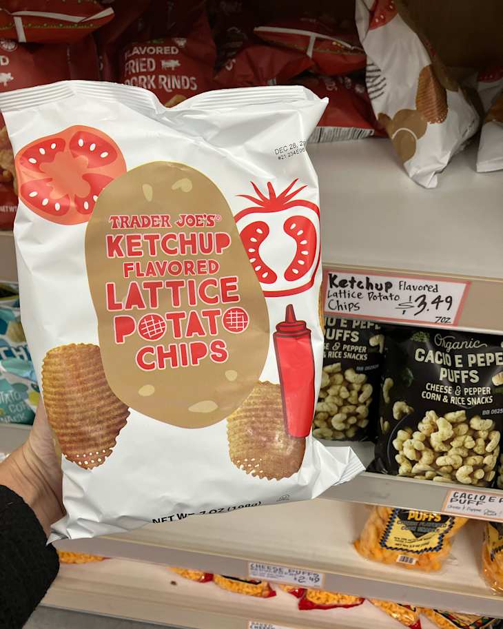 Ketchup flavored lattice potato chips in a white bag held in a grocery aisle, with other snacks in the background.
