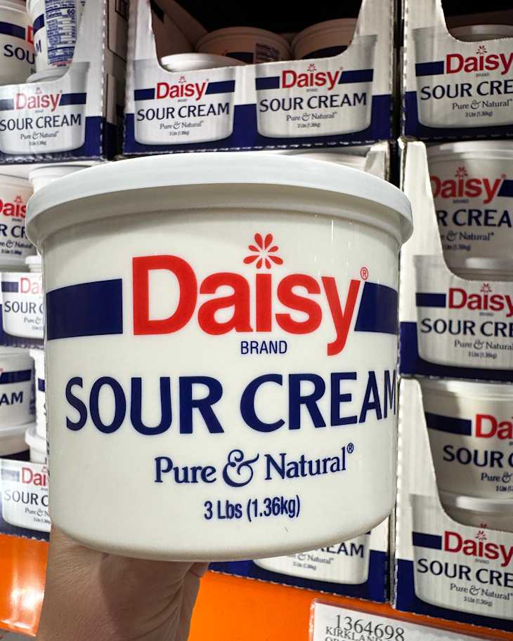 Large container of Daisy brand sour cream, labeled "Pure & Natural," held in a hand with more containers in the background.