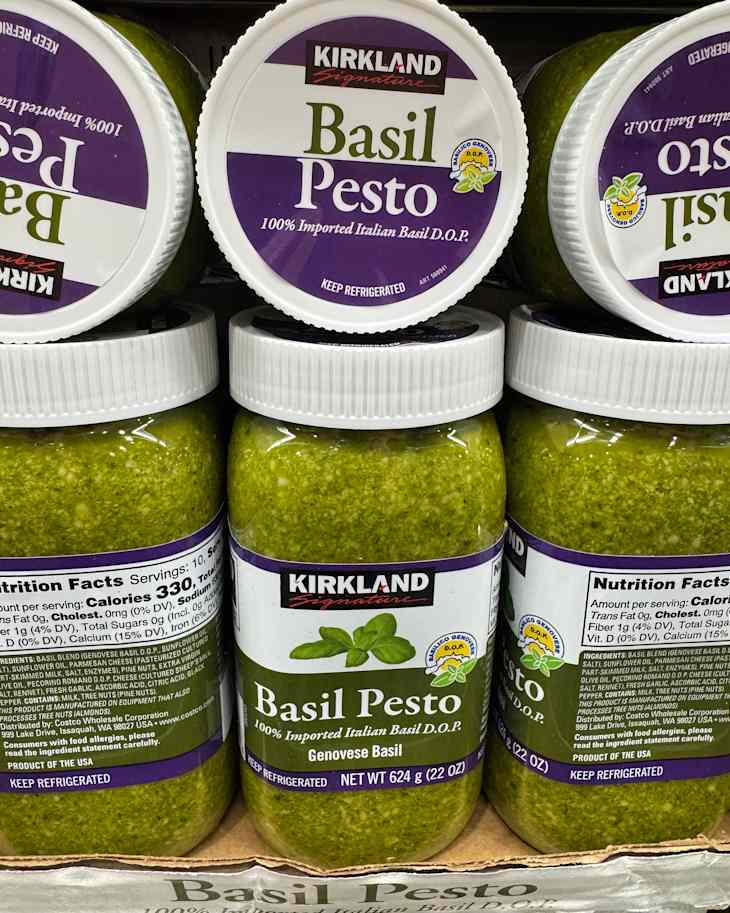 Jars of Kirkland Signature Basil Pesto stacked on a shelf, featuring green pesto and nutrition labels.