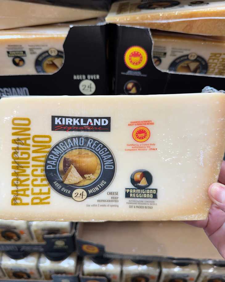 Block of Parmigiano Reggiano cheese labeled "Kirkland Signature," aged over 24 months, held in a hand.