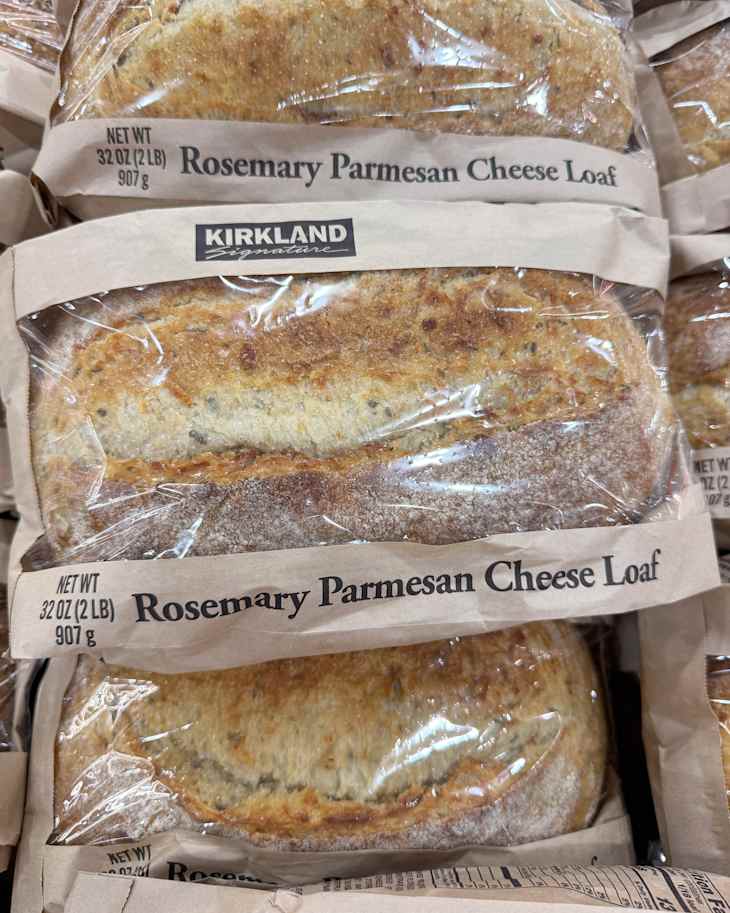 Rosemary Parmesan Cheese Loaves in clear packaging stacked on a shelf, labeled with weight and brand.