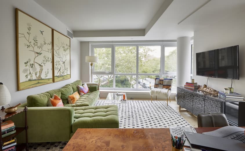 Living area with white walls and large windows, daylight coming in. Left wall has green velvet sofa with colorful pillows. Large illustrated diptych on wall above the sofa. Right wall has a long media console with TV mounted on wall above. Black and white patterned rug on floor.