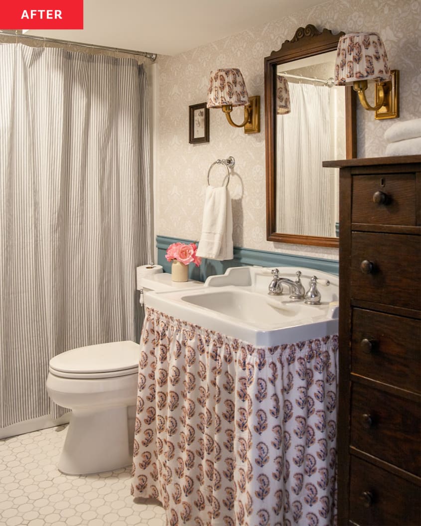 A finished bathroom with a shower hidden by a striped shower curtain on the left side, and in the center, a vanity that has a sink skirt. The floral pattern on the skirt matches the sconces on either side of a wood mirror, and there's a wood dresser beside the sink on the right. A toilet is in between the sink and shower.