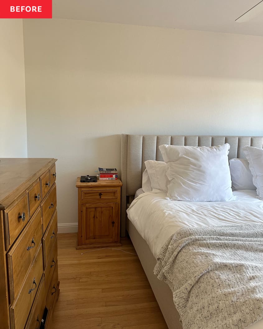 Primary bedroom before renovation: White walls with no art or decoration, wood dresser against wall with a plant, candles, bed with quilted gray headboard, sliding glass doors letting in light with open pale neutral curtains