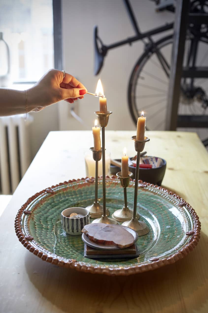 在餐桌上的一个陶瓷装饰碗里，用手点燃一支黄铜烛台上的蜡烛，旁边还有两个烛台