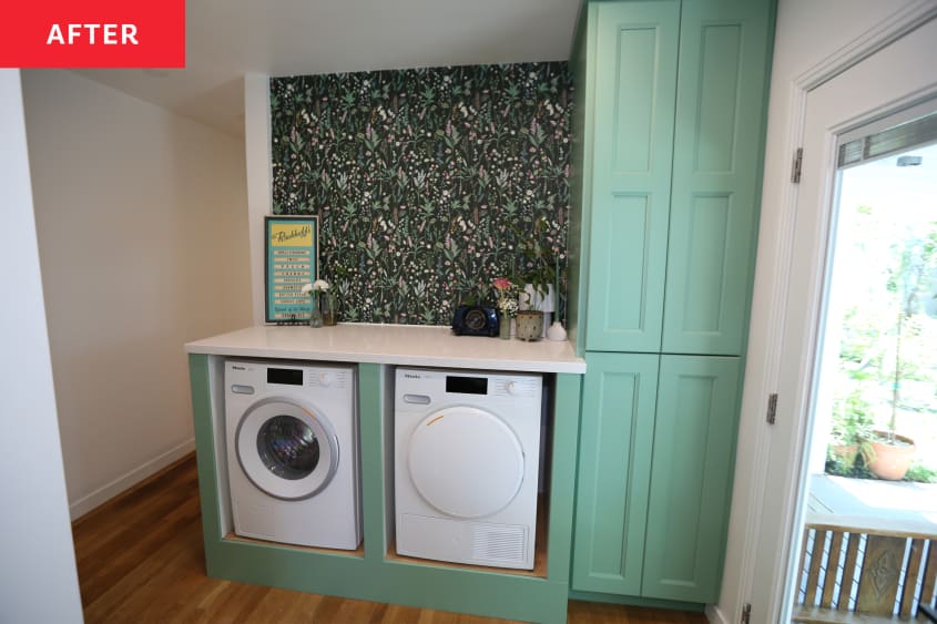 Washer/ dryer appliances sit under white stone countertops with dark colored botanical wallpaper on wall behind. Large green cabinet on side of appliances.