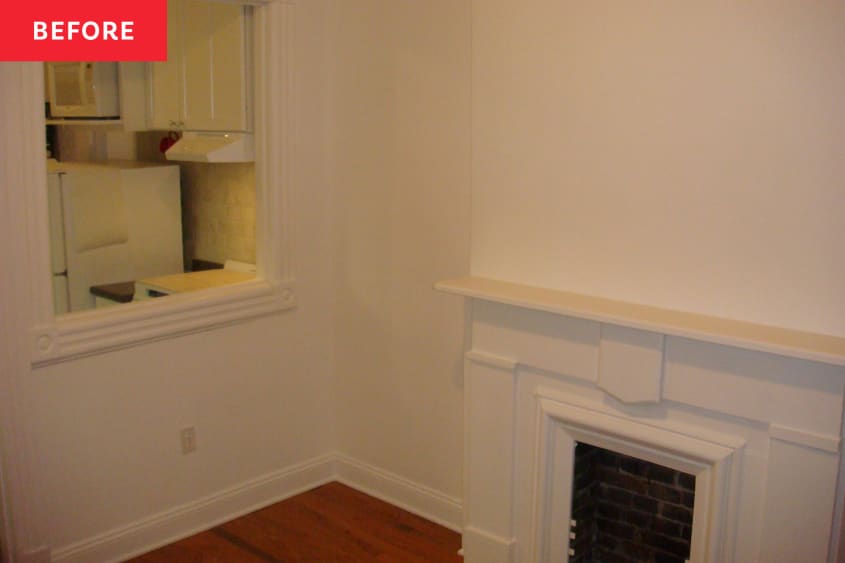 Fireplace in empty white living room with cut out window peeking into kitchen.