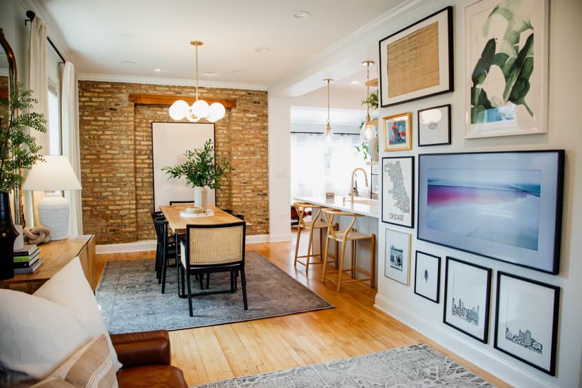Brick walled dining room with wooden rectangular dining table surrounded by black framed dining chairs. Art print gallery wall on living room wall.