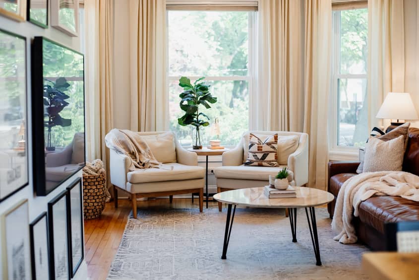Two neutral colored arm chairs near curtain lined window in light filled living room.
