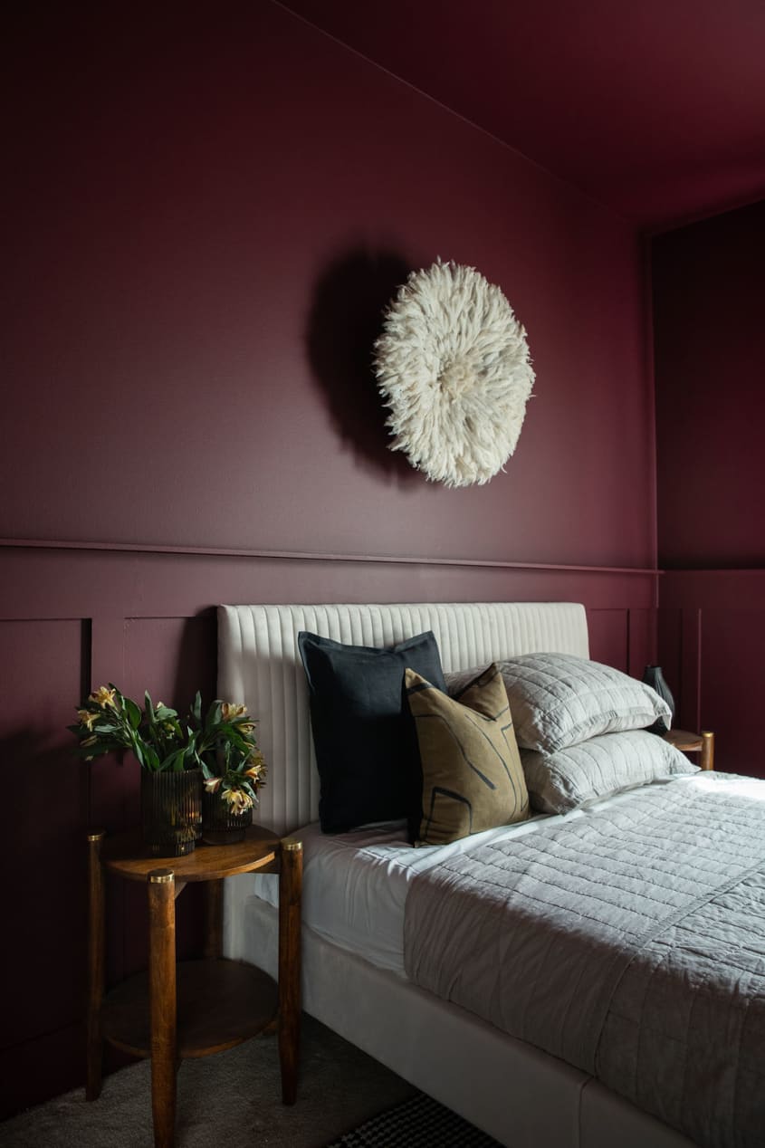 Beautiful moody burgundy bedroom with a white juju hung above the cream headboard and light bedding on the bed with a wooden side table as a nightstand