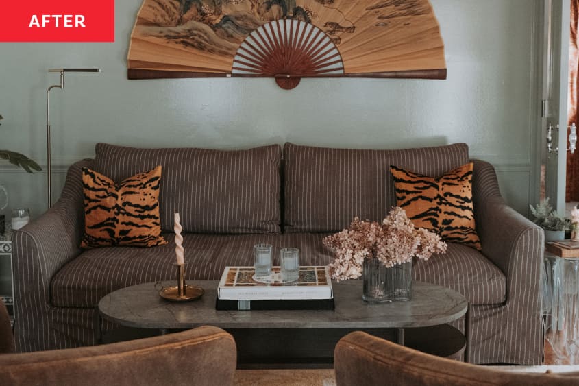 Photo after home office remodel: brown loveseat with white stripes, tiger throw pillows, large decorative fan on wall above, marble/stone coffee table with dried flower arrangement, books, candle