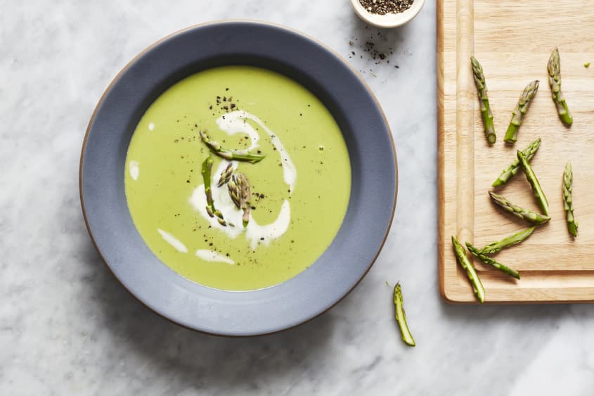 Bowl of asparagus soup sits on the counter next to a cutting board with cut ends of asparagus.