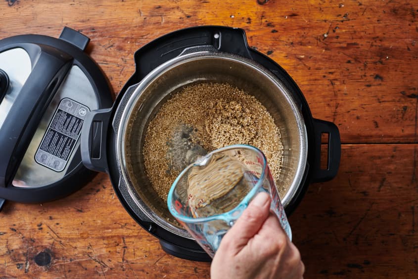 A glass liquid measuring cup full of water is added to the uncooked quinoa, in the instant pot.