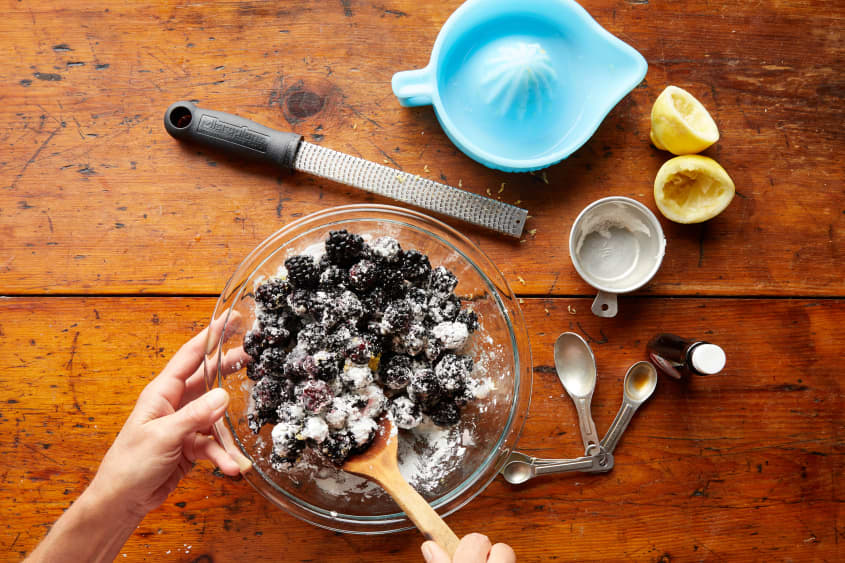 A Hand tosses sugar, lemon, berries, and extract together.