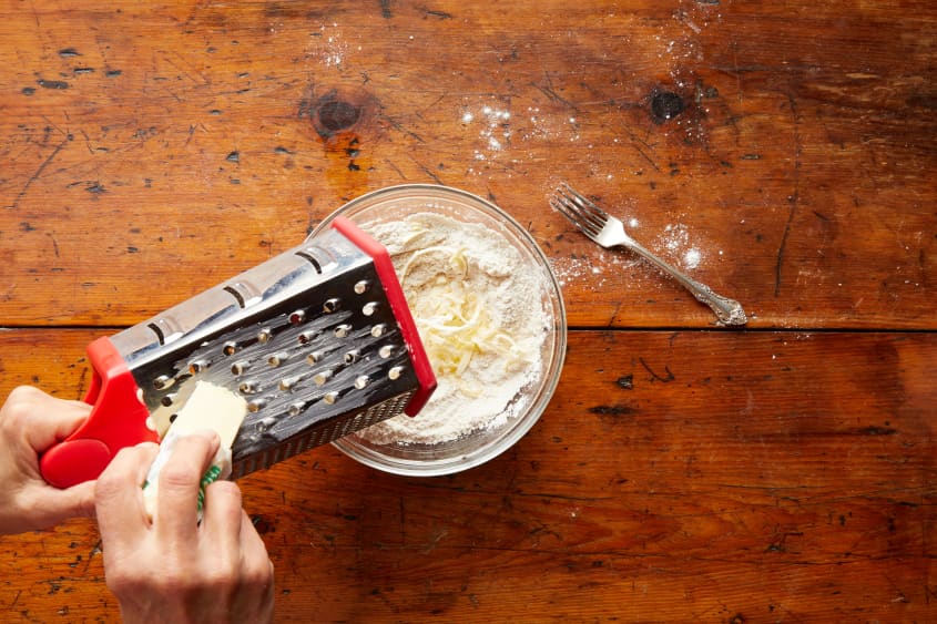 With a box grater, a hand grates butter into the dry ingredients, in a bowl.