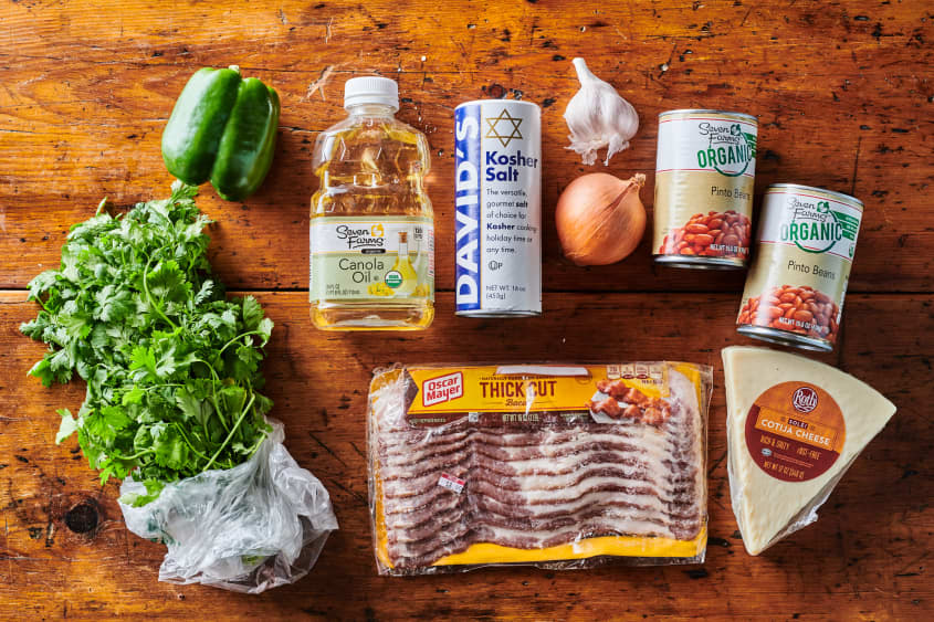 Refried beans ingredient line up; includes two cans of pinto beans, cotija cheese, thick cut bacon, cilantro, green bell pepper, canola oil, kosher salt, yellow onion and garlic