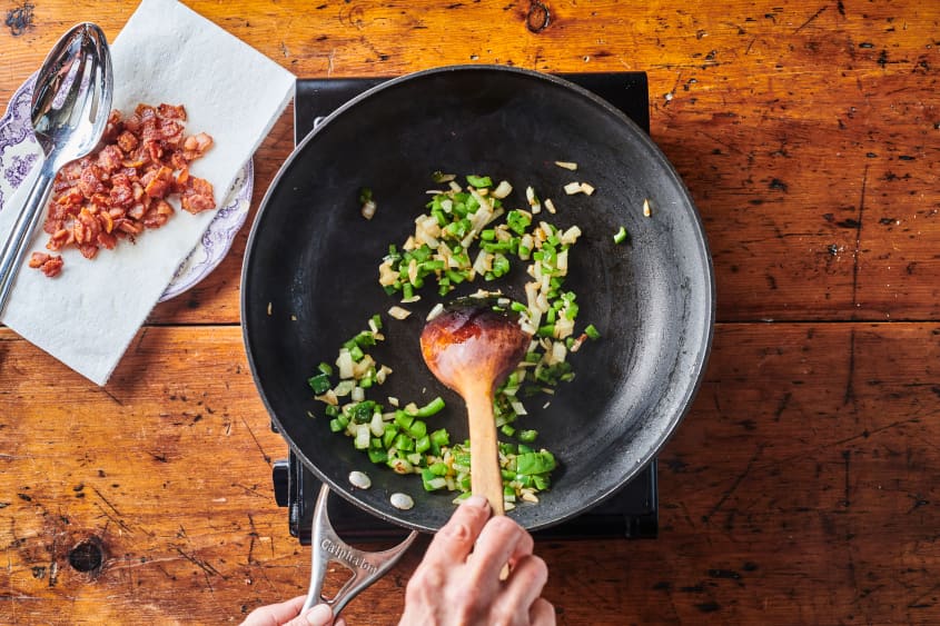 Skillet with chopped green bell peppers and onions with cooked bacon set aside on the counter