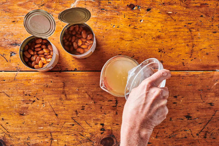 Two opened cans of pinto beans with someone pouring bean liquid into glass measuring cup